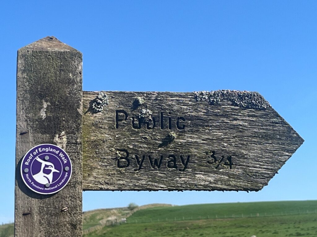 A wooden finger post against a blue sky points right along a Public Byway. On the upright post there is a round waymarker disc with a white outline of a bird's head. Around the bird is the following writing - Roof of England Walk, North Pennines National Landscape.