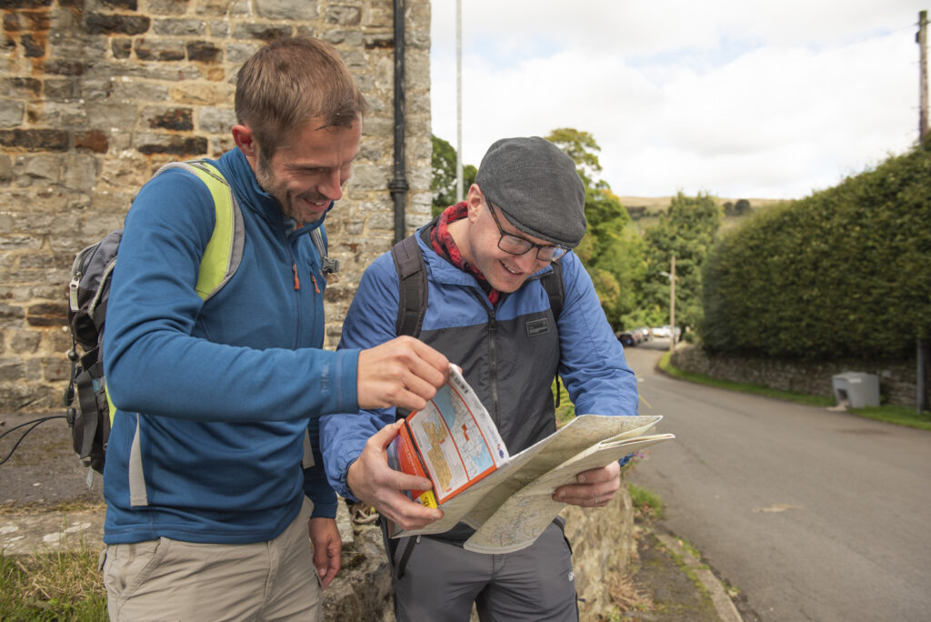 Two smiling male walkers studying a paper map near a stone wall on a village road, both dressed in outdoor gear and carrying rucksacks.