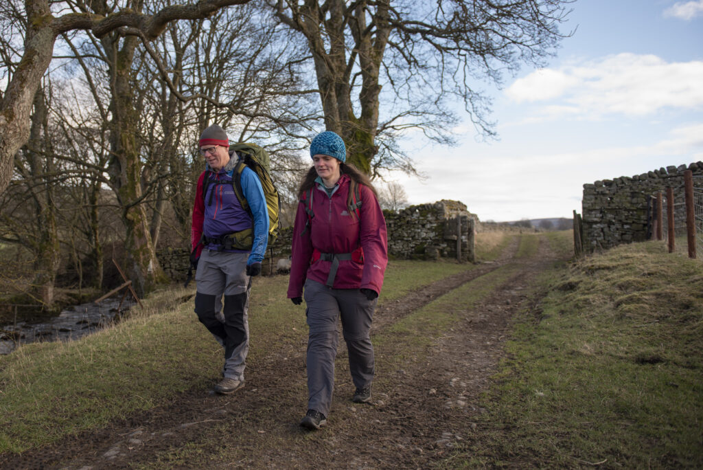 Two walkers walking along a rural path lined by leafless trees and stone walls, with a small stream to one side. They appear to be mid-conversation and are dressed in winter walking gear.